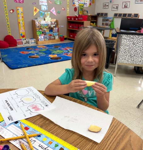 A young girl sits at a desk holding an apple slice. A colorful rug, pencils, and paper are on the table.