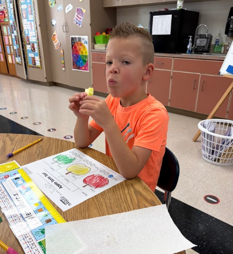A child in an orange shirt sits at a table, holding an apple slice. A worksheet with apple images and text sits before him.