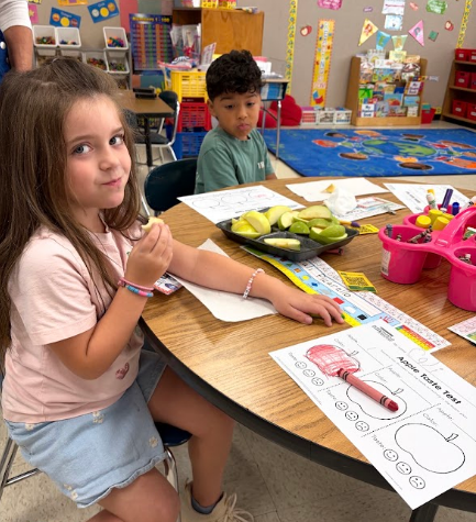 A young girl eats while sitting at a table with educational materials. A boy is seated behind her.