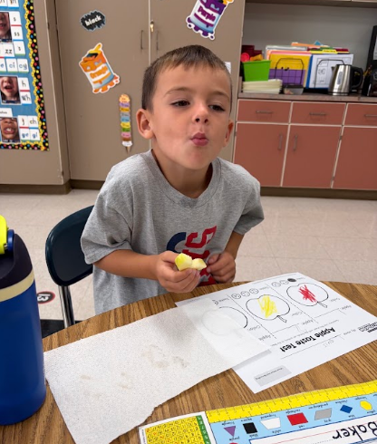 A child sits at a desk, holding food, and looking up. In front of him is a worksheet.