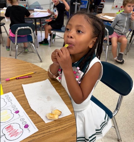 Child in a classroom eating an apple. Behind her, other children are seated at tables.