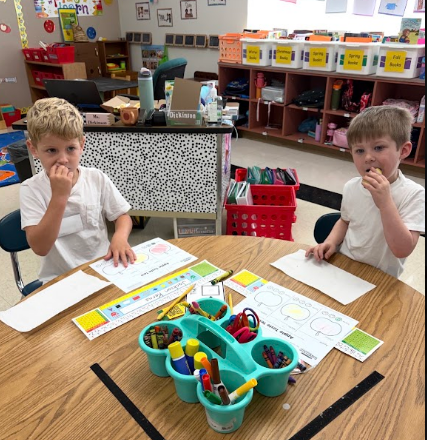 Two children sit at a desk, one eating, the other with a finger to their lips. Various papers, markers, and a ruler are on the desk.