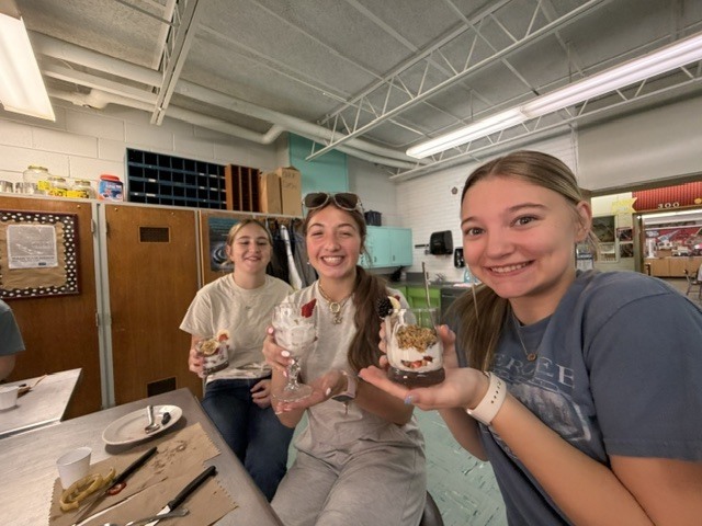 Three women holding dessert glasses in a kitchen with wooden cabinets, countertops, and fluorescent lights.