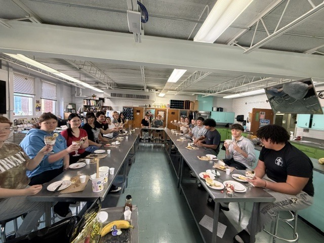 A classroom with several people sitting at long tables, facing each other, with food and drinks on the tables.