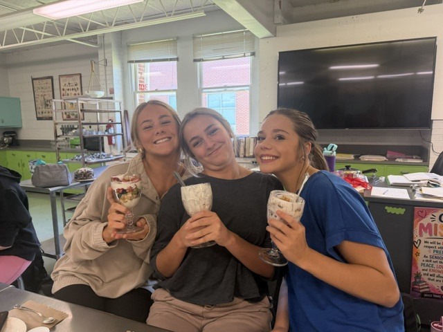Three women hold milkshakes. They sit around a table in a kitchen. A flat screen TV is behind them.