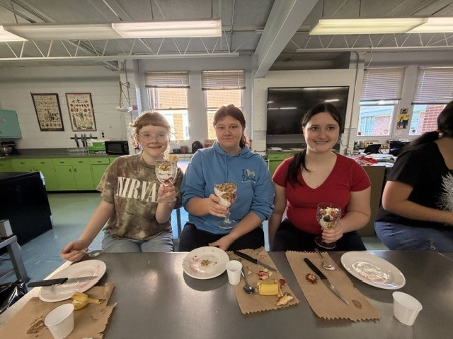 Three people sit at a table with plates, cups, and knives, holding drinks and smiling. Behind them is a kitchen with cabinets and windows.