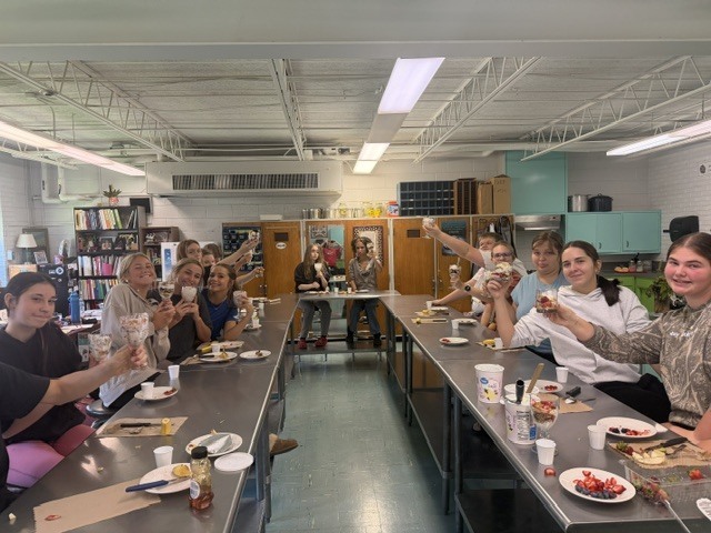 People sitting at tables in a room with raised hands, plates, and cups; some are smiling.