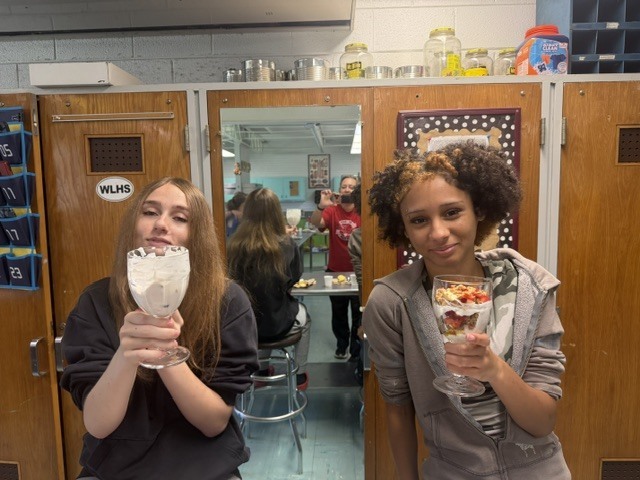 Two women hold glasses of dessert in a room. A mirror and people are behind them.