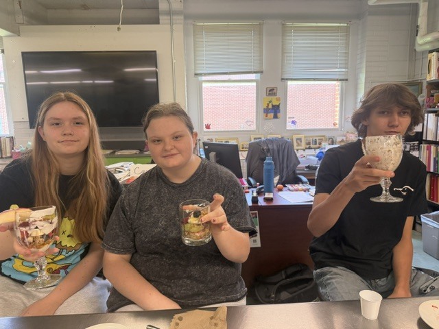 Three students in a classroom hold drinks. One has a dessert in a tall glass.