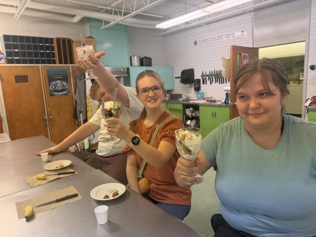 Two people in a kitchen holding up dessert cups. One wears a watch and orange shirt.