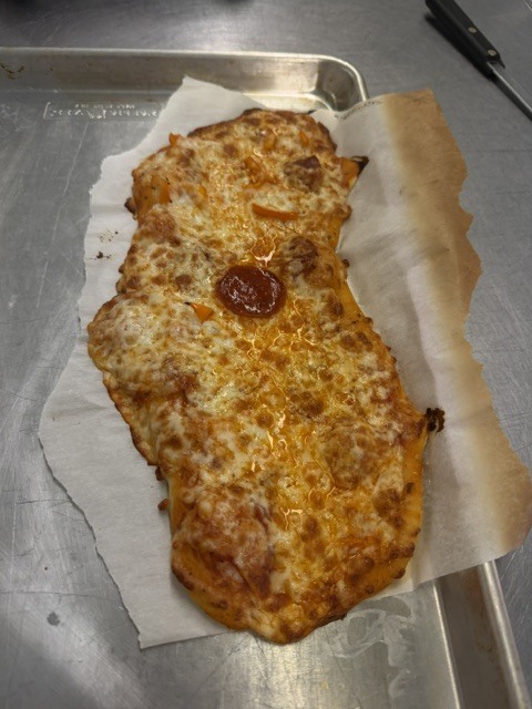 Pizza on a tray, with cheese and pepperoni, on a piece of paper. A black handle is visible.