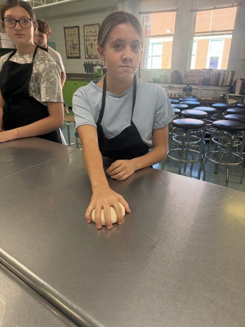 Two girls in aprons, one kneading dough, in a classroom with stools, windows, and framed pictures.
