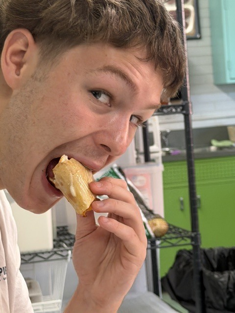 A man bites a piece of food with his mouth open, showing surprise. He is in a kitchen with shelves and cabinets.