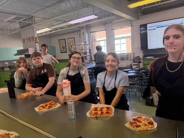 Five people in aprons stand in a kitchen, posing for a photo. They are in front of a table with pizzas.