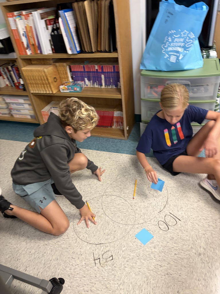 Two children sit on a classroom floor, drawing circles and numbers. A bookshelf and a blue bag are in the background.