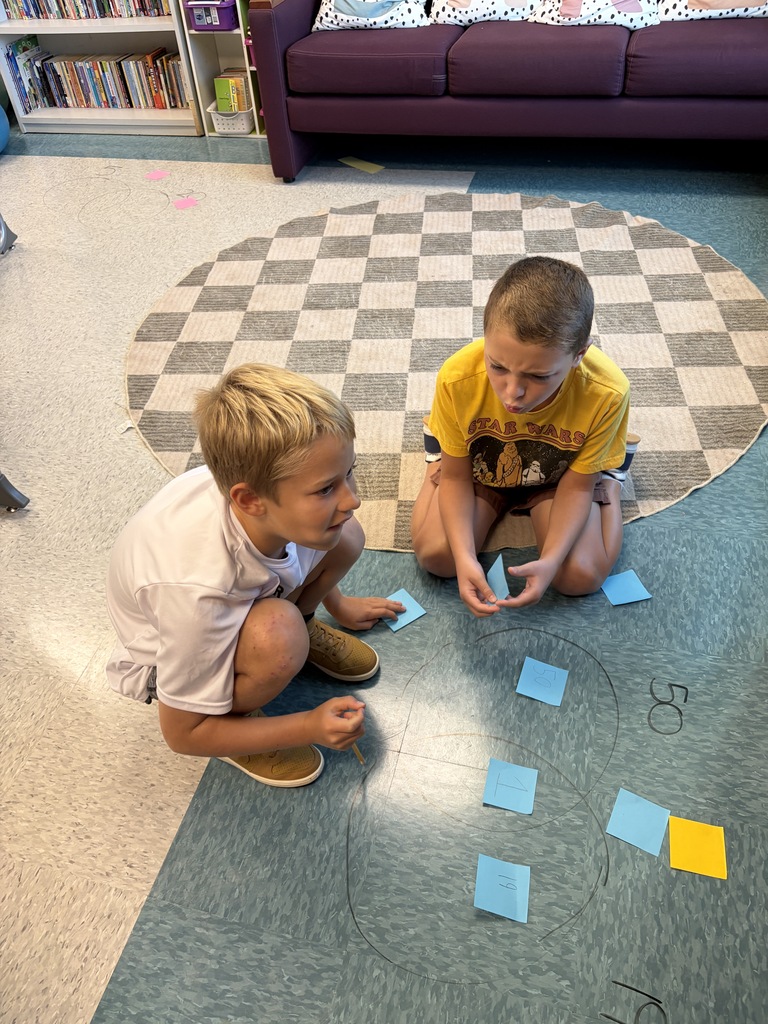 Two boys kneel on a carpeted floor, each holding cards with numbers. A rug with a checkered pattern is behind them.
