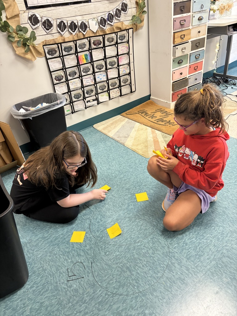Two girls in glasses on a blue floor, one using a yellow card, the other using scissors.