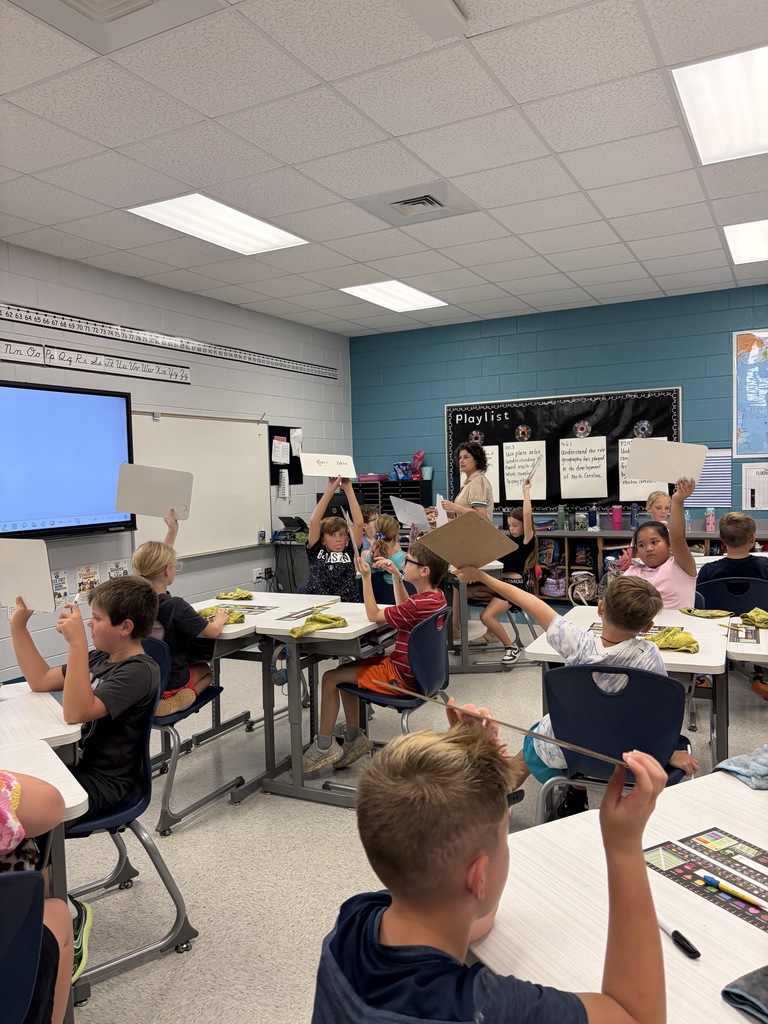 Children seated at tables in a classroom raise white cards with hands up. A teacher stands at the front.