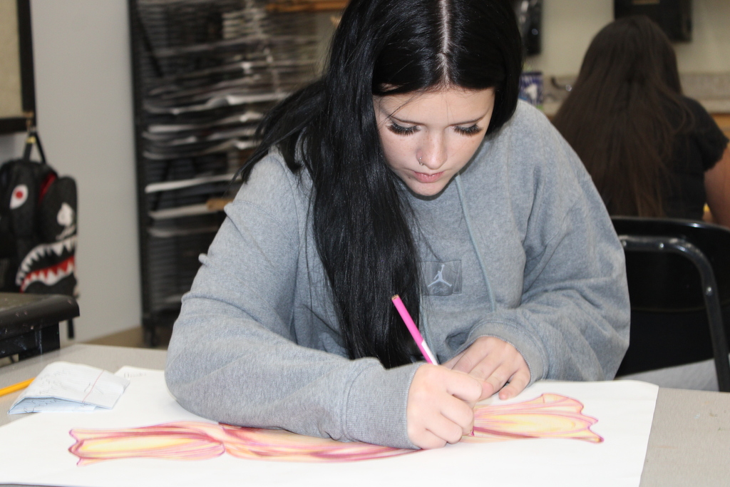 Person in gray sweatshirt drawing on white paper with a pink pen. Another person sits in the background.