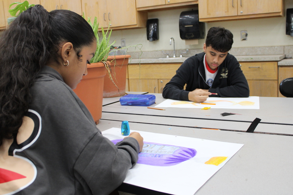 Two students sit at a table, drawing on large sheets of paper with markers. One holds a plant in a pot.
