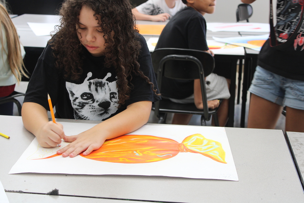 A young person with curly hair colors an orange fish on a large white sheet of paper.