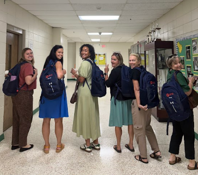 Six people in a hallway with backpacks, wearing summer clothes, and posing for a picture.