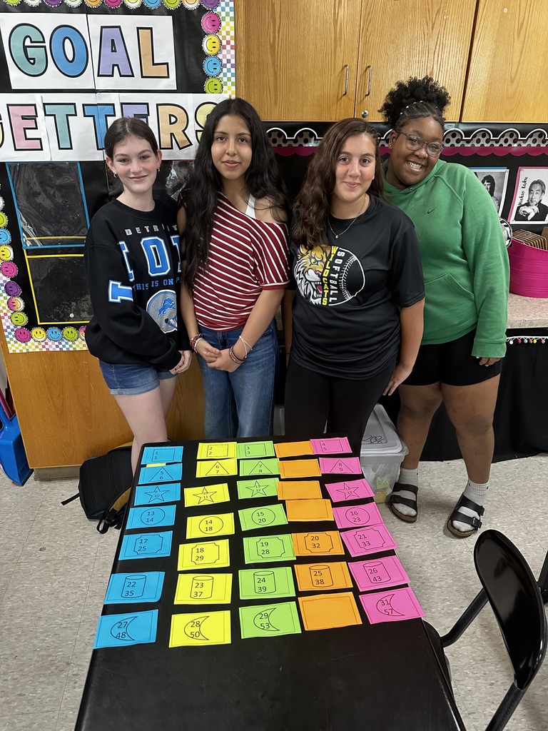 Four students stand in a room, near a table covered with colorful, labeled squares. Behind them, a wall displays a poster with "Goal Setters" text.