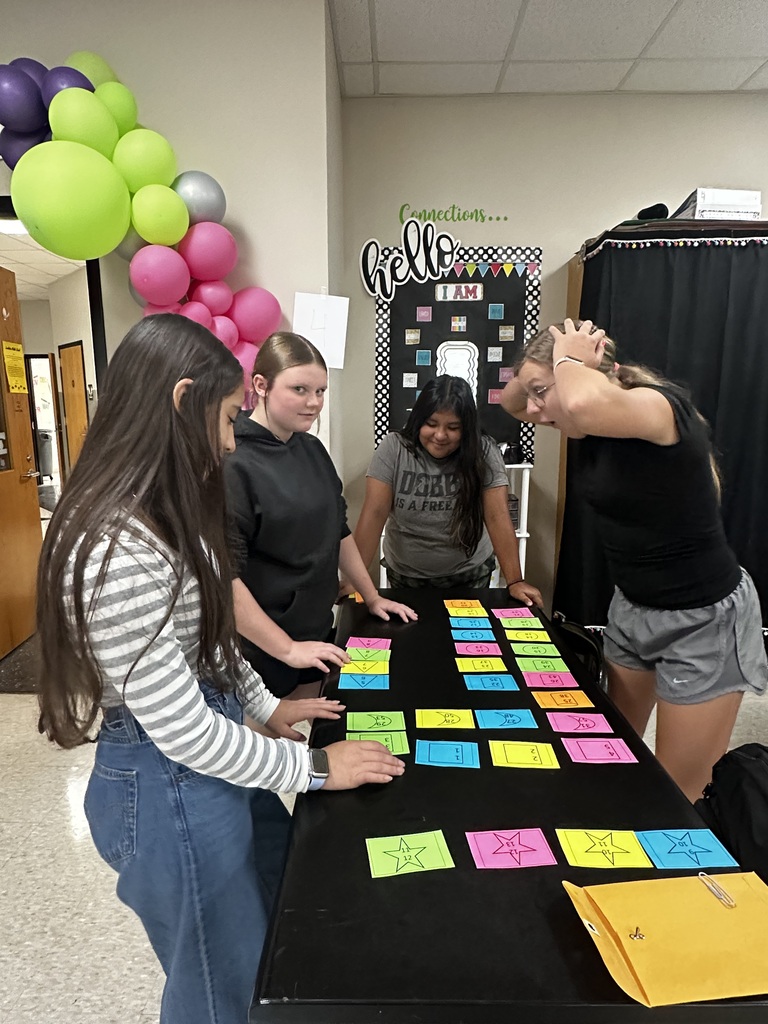 Five people at a table with colorful sticky notes. A bulletin board reads "Hello". Behind them, balloons hang on the wall.