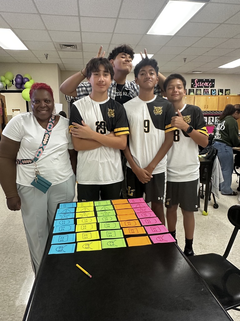 Five individuals, two adults and three children, stand near a table with multicolored squares. They are in a room with ceiling lights.