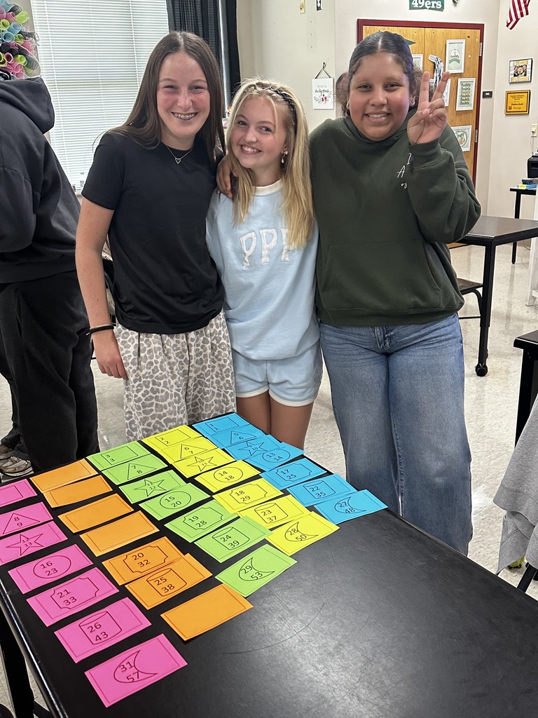 Three young women stand beside a table with colorful, square cards. A few others are in the background.