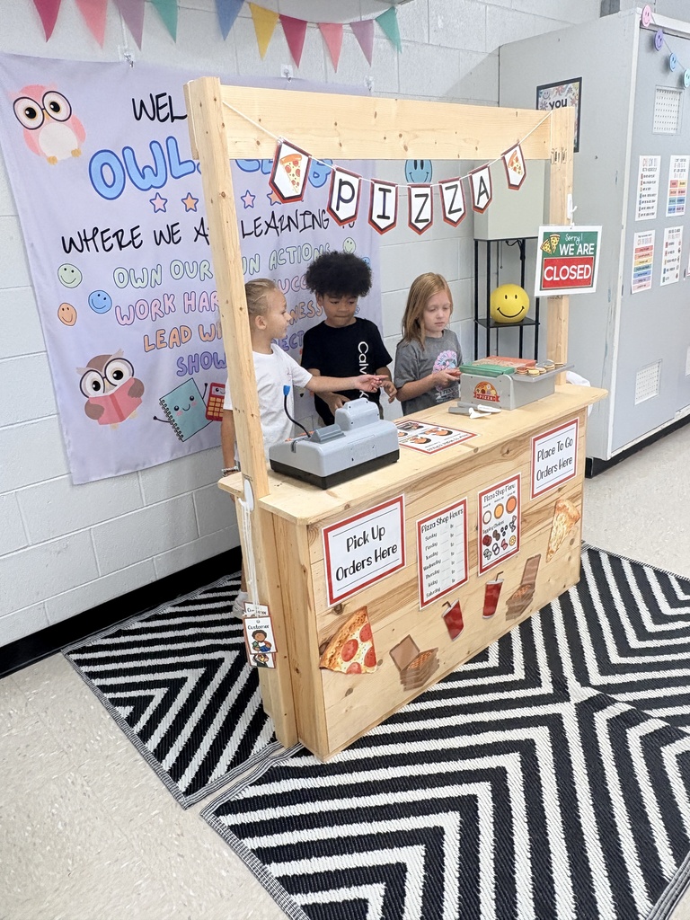 Three children stand behind a wooden counter with a menu, cash register, and small items. A black-and-white rug lies on the floor.
