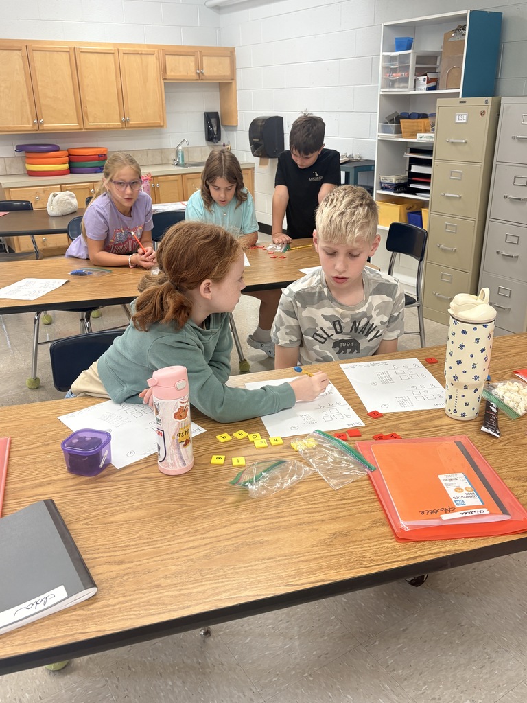 A classroom with children at desks. Two children sit at a table with papers and a pink cup.