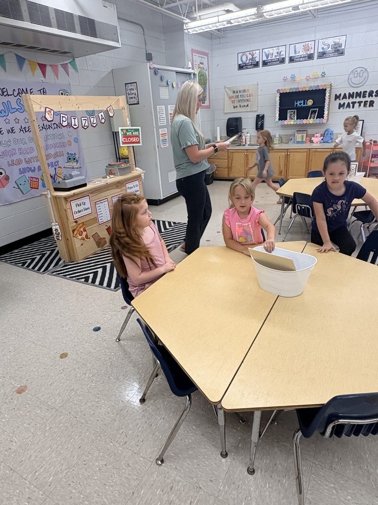 Four young children sit at a table in a classroom. A teacher stands behind them. Bright posters adorn the walls.