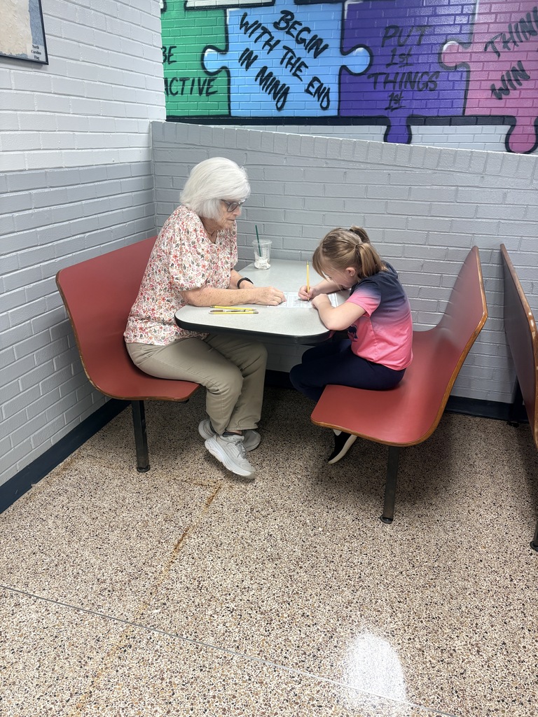 Two people sit at a table, with one older and the other younger, both facing a piece of paper.