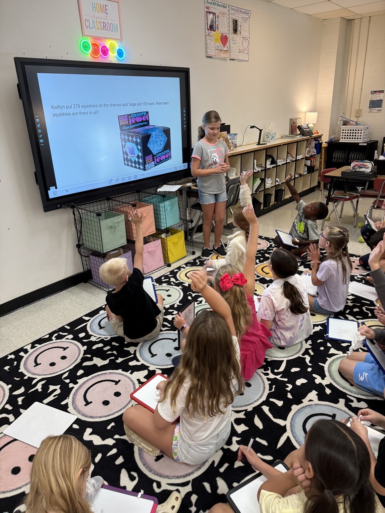 A classroom with a large screen and a teacher. Children sit on the floor with raised hands.