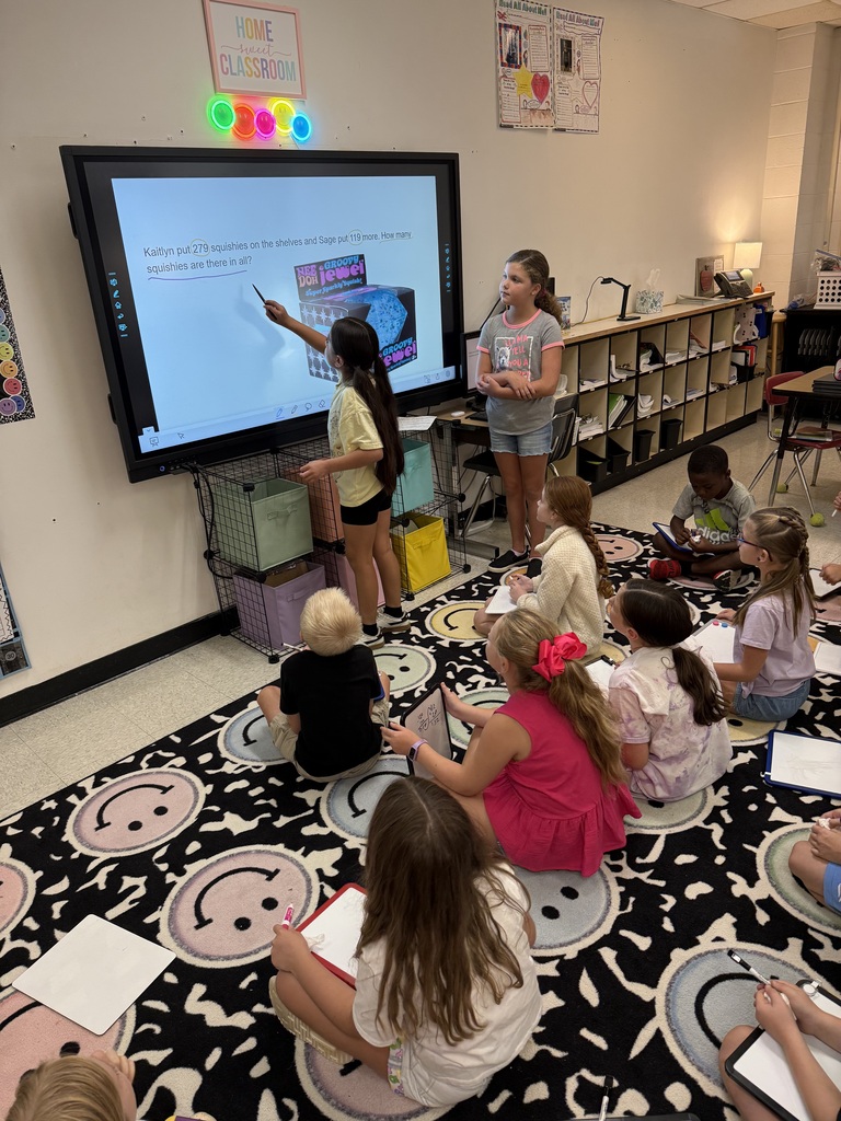 A classroom with a teacher at a screen. Several children are seated on the floor in front of her.
