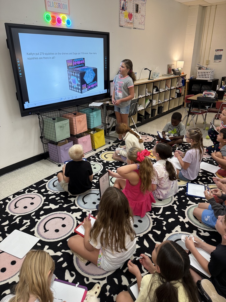 A classroom with a large screen; a teacher stands before it while a group of students sit on the floor.