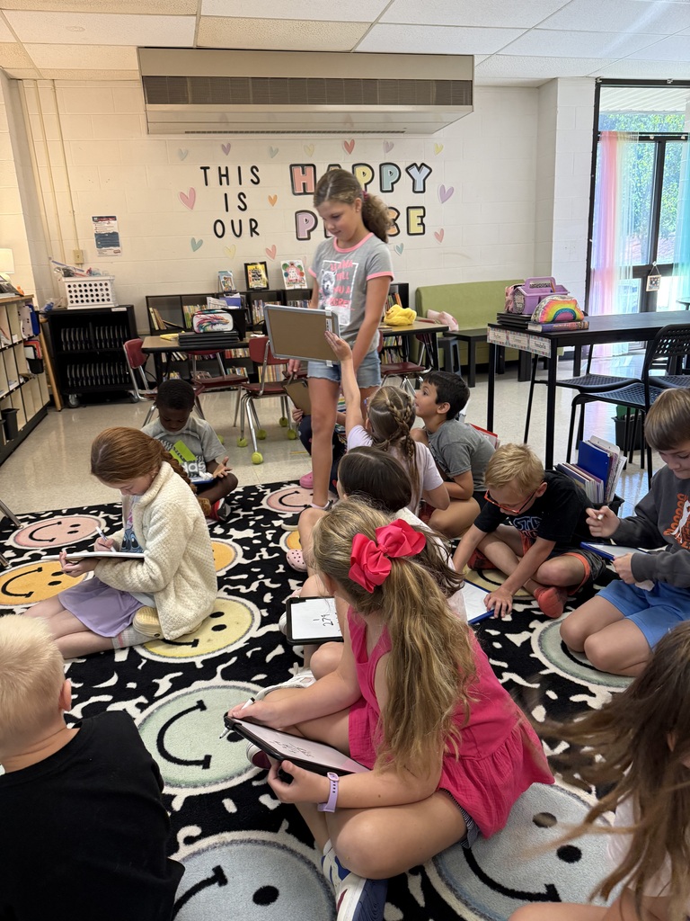 Kids in a classroom, seated in a circle, listening to a girl holding a laptop. Bright, colorful classroom with tables and bookshelves in the background.