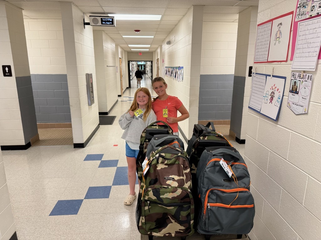 Two people stand in a school hallway with several bags, including camouflage and orange backpacks, while papers are pinned to a wall.