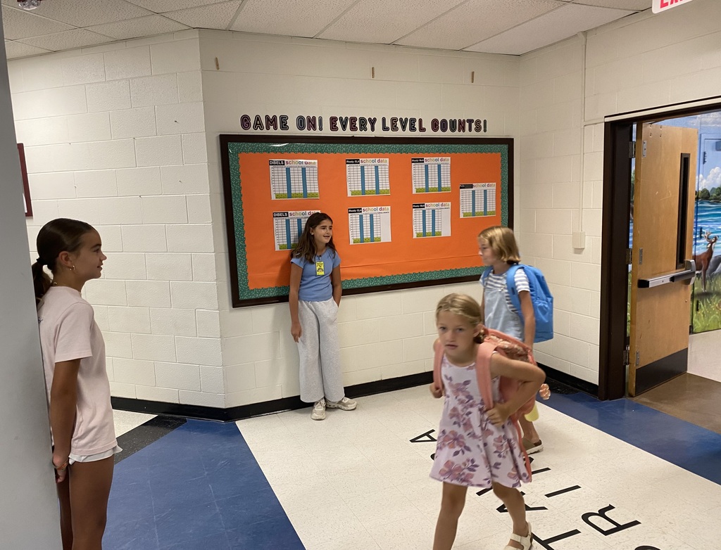 Four young girls in a hallway with one girl carrying a backpack. A colorful board is on the wall.
