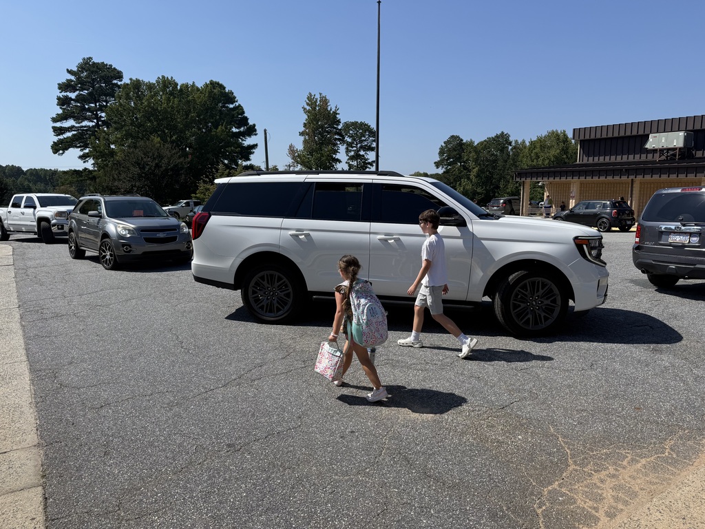 Two children cross a parking lot in front of a large SUV. Behind them, a business building stands.
