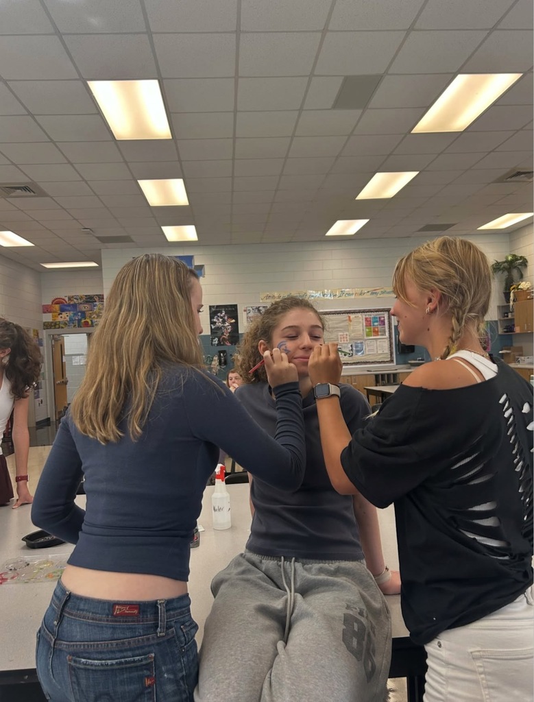 Three women in a classroom. One has her eyes closed as another puts makeup on her face.