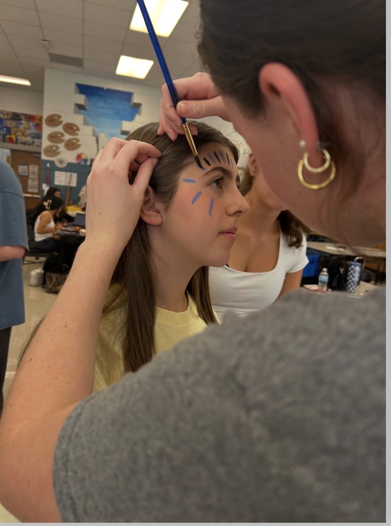 A woman paints blue paint on another woman's face in a room. Other people are in the background.