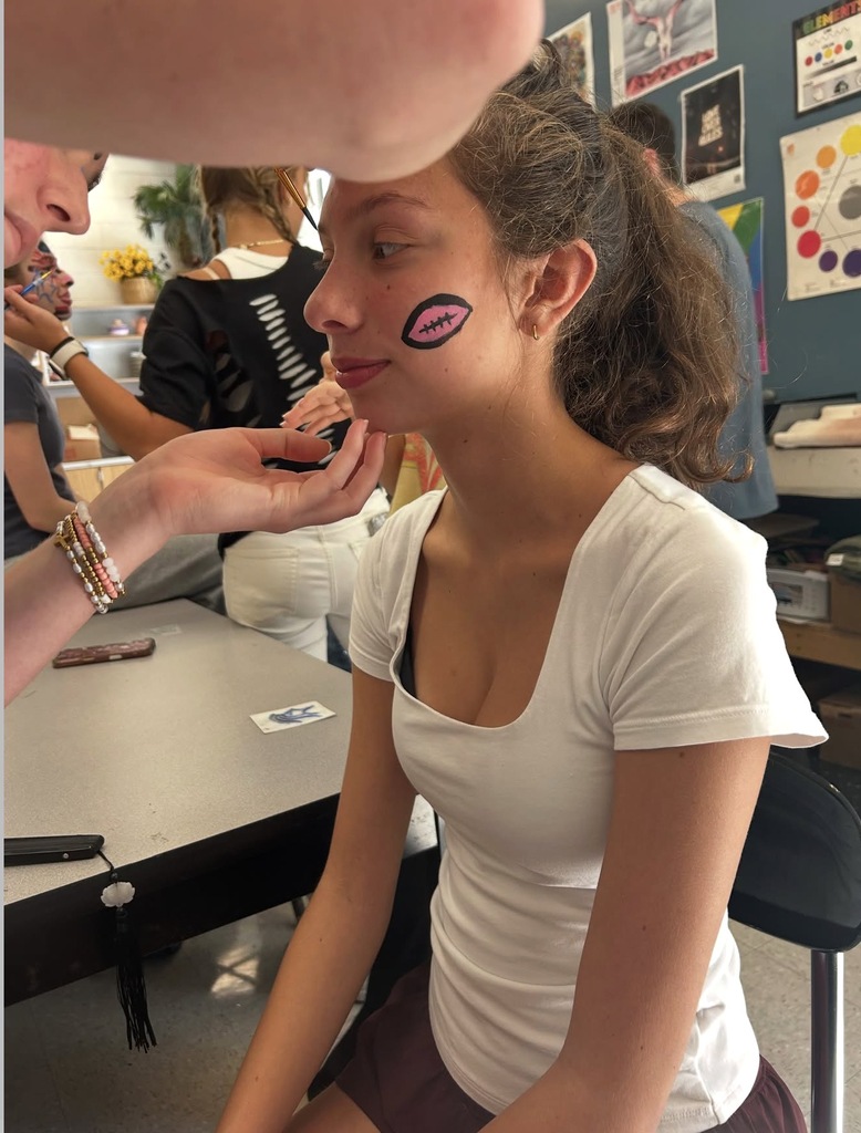 A girl with face paint sits on a chair as a person with a phone paints her face.