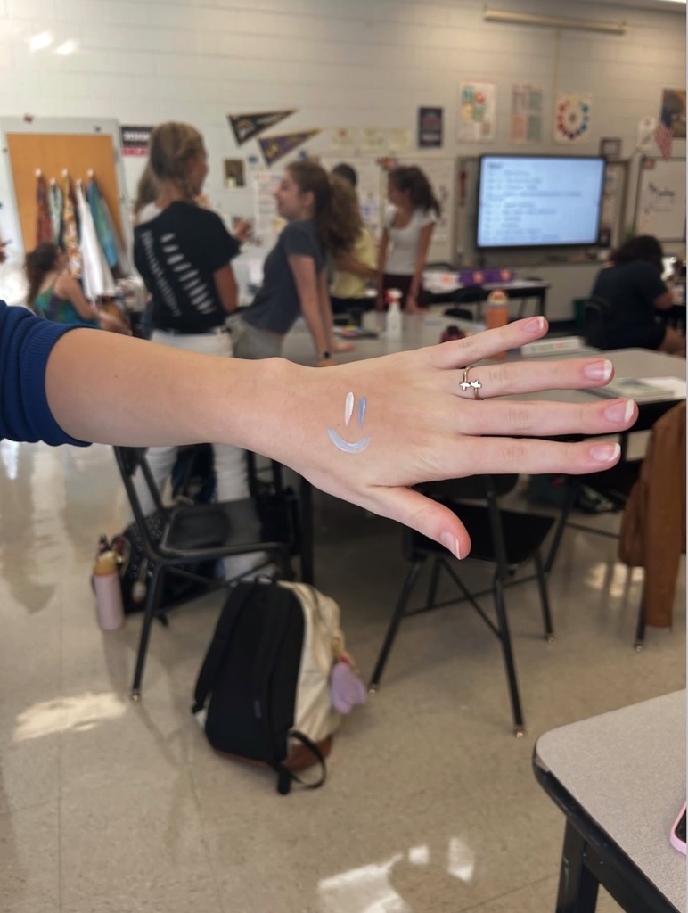 A hand with white paint on it. In the background, a group of students are working at tables.