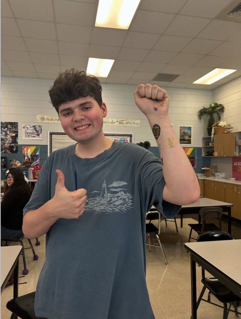 Person with a thumb up and a smile standing in a classroom with tables and chairs.