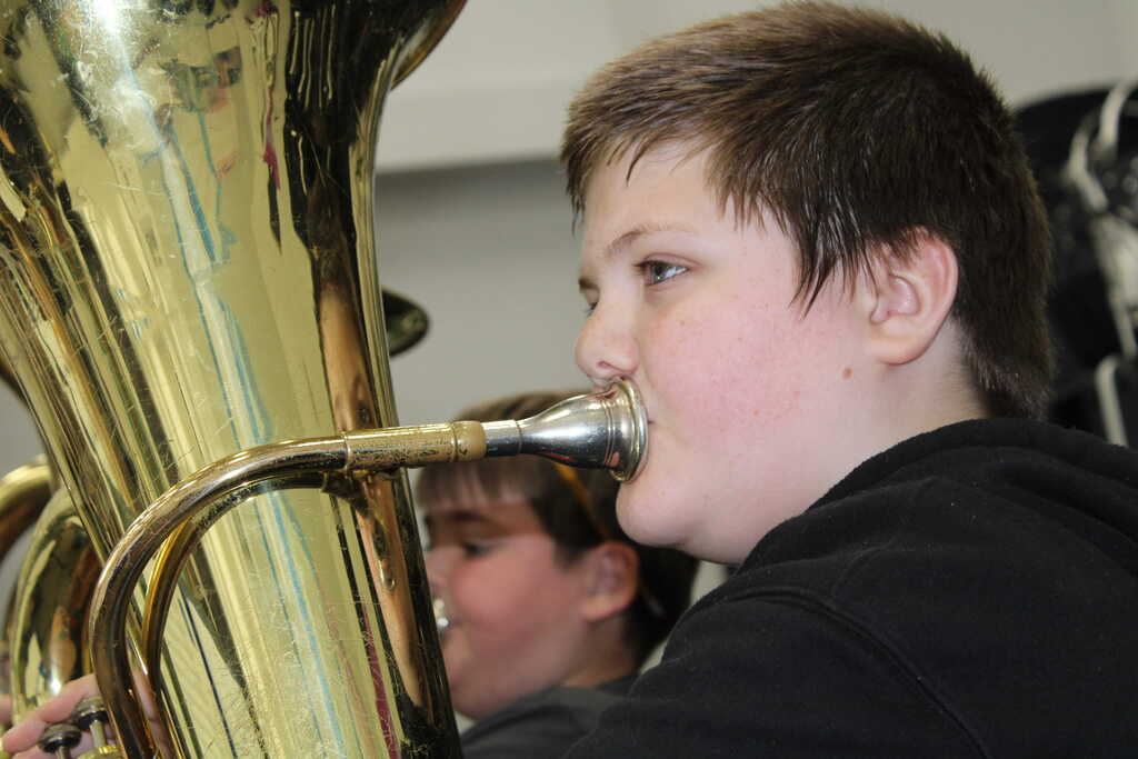 A boy plays a brass tuba, focused on his performance with another boy visible in the background.