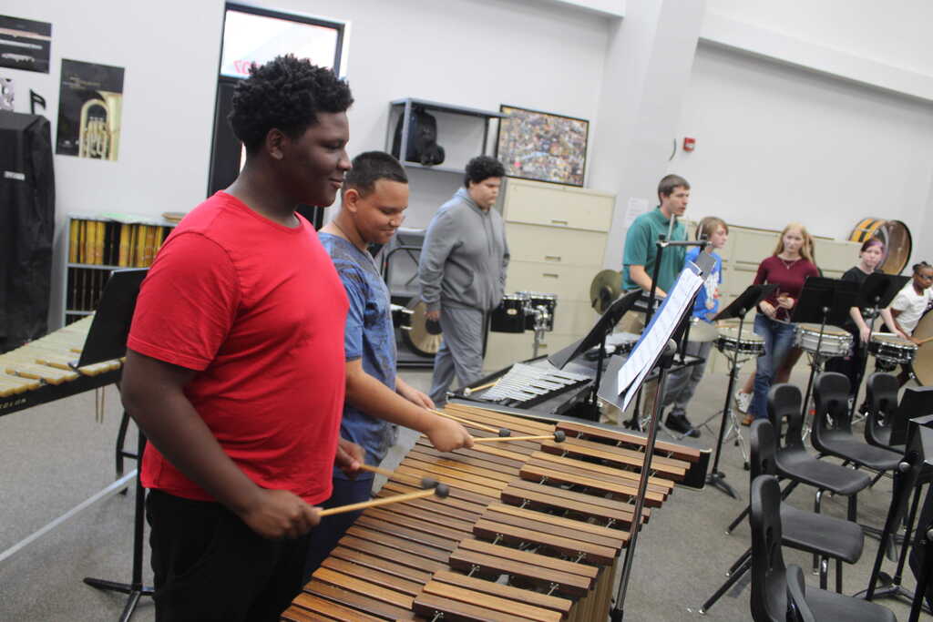 A group of students in a music room, one playing xylophone, others standing and looking at sheet music.