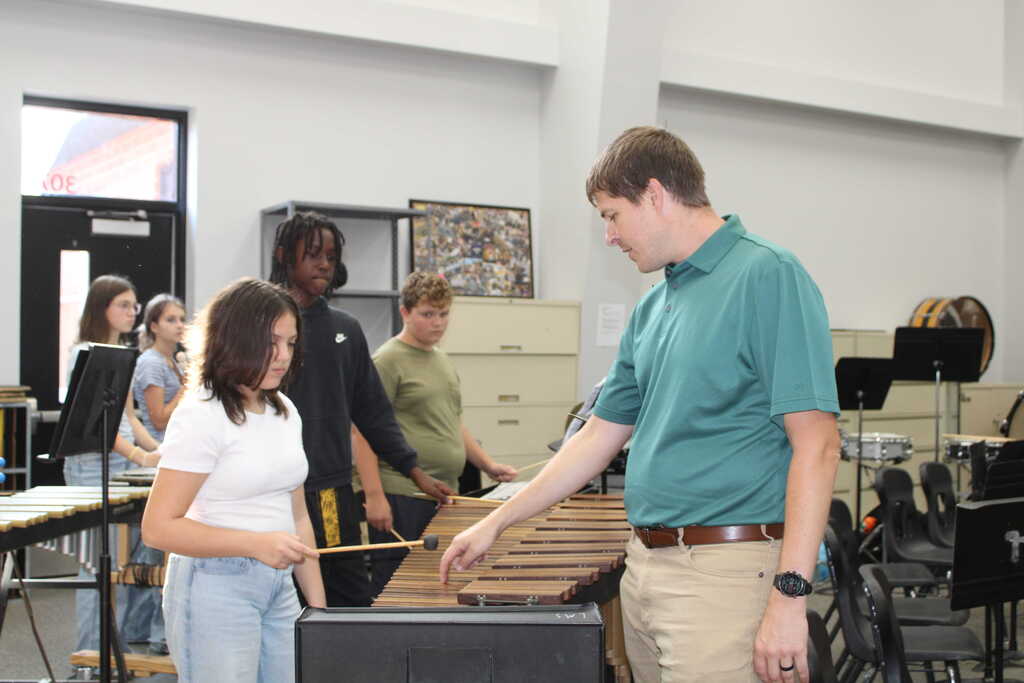 Several students in a music room, one holding drumsticks, another playing xylophone. A teacher assists.