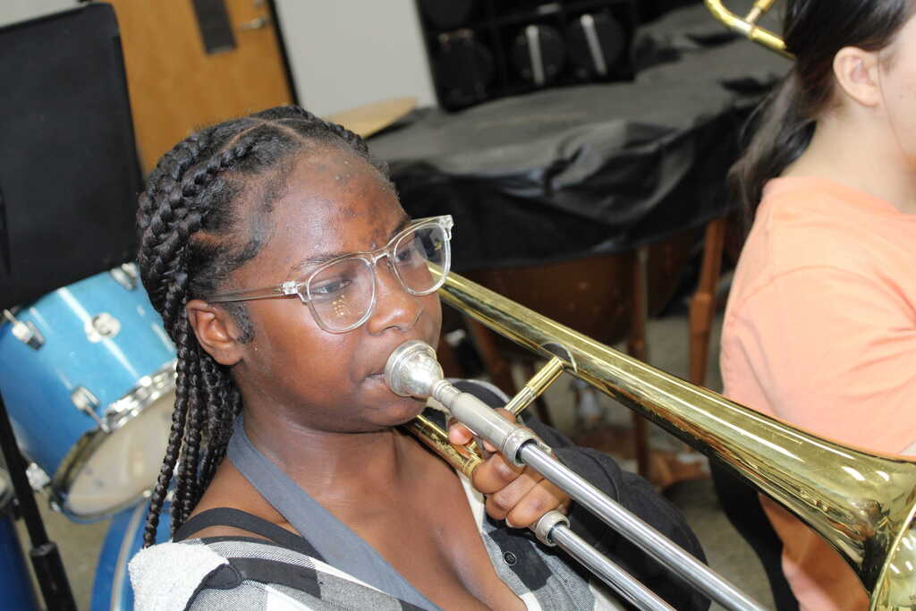 A girl with braided hair wearing glasses plays a trombone while another girl in an orange shirt looks on.